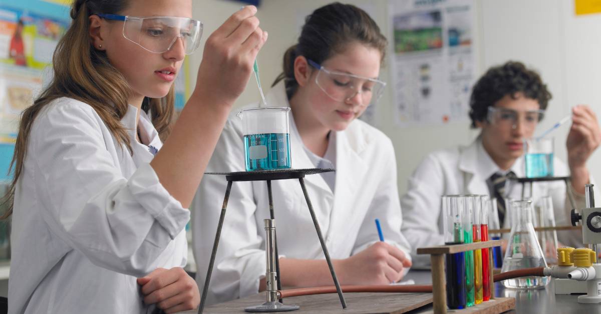 Three students in a chemistry class wearing laboratory coats and glasses, taking notes and experimenting with beakers.