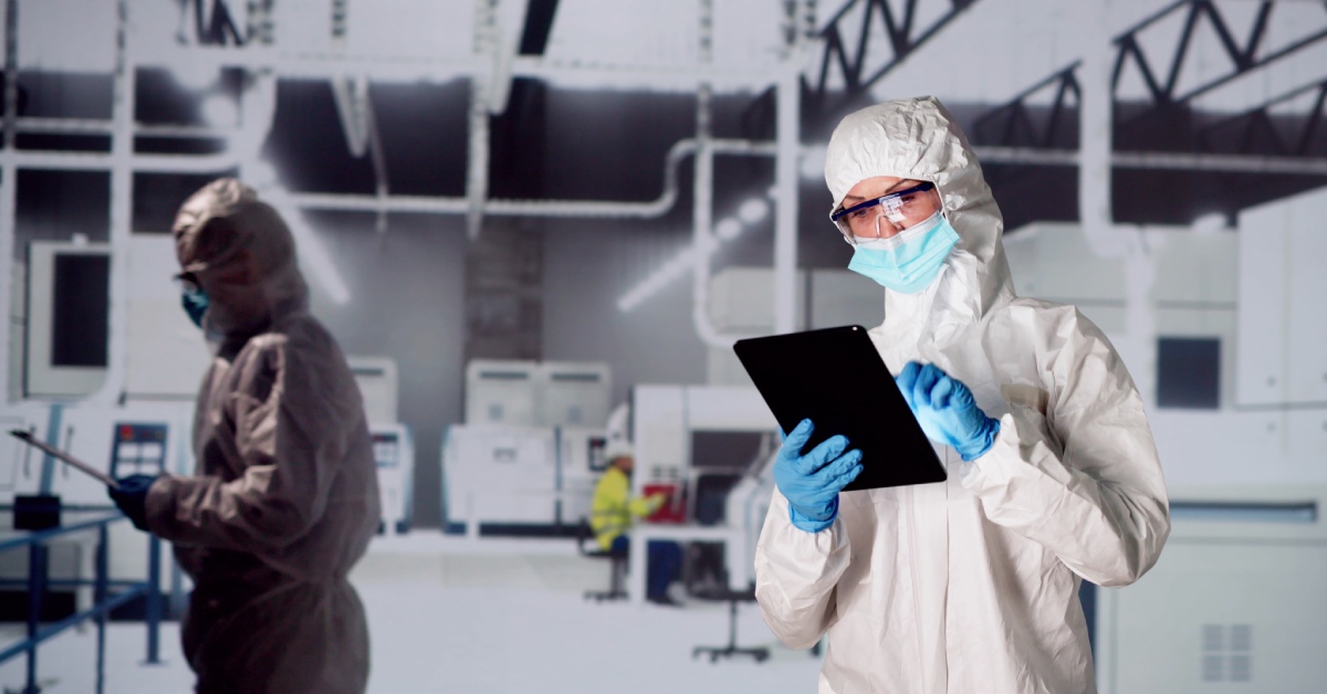 Two people inside a facility wearing laboratory suits, gloves, and eyeglasses and taking notes on a tablet.