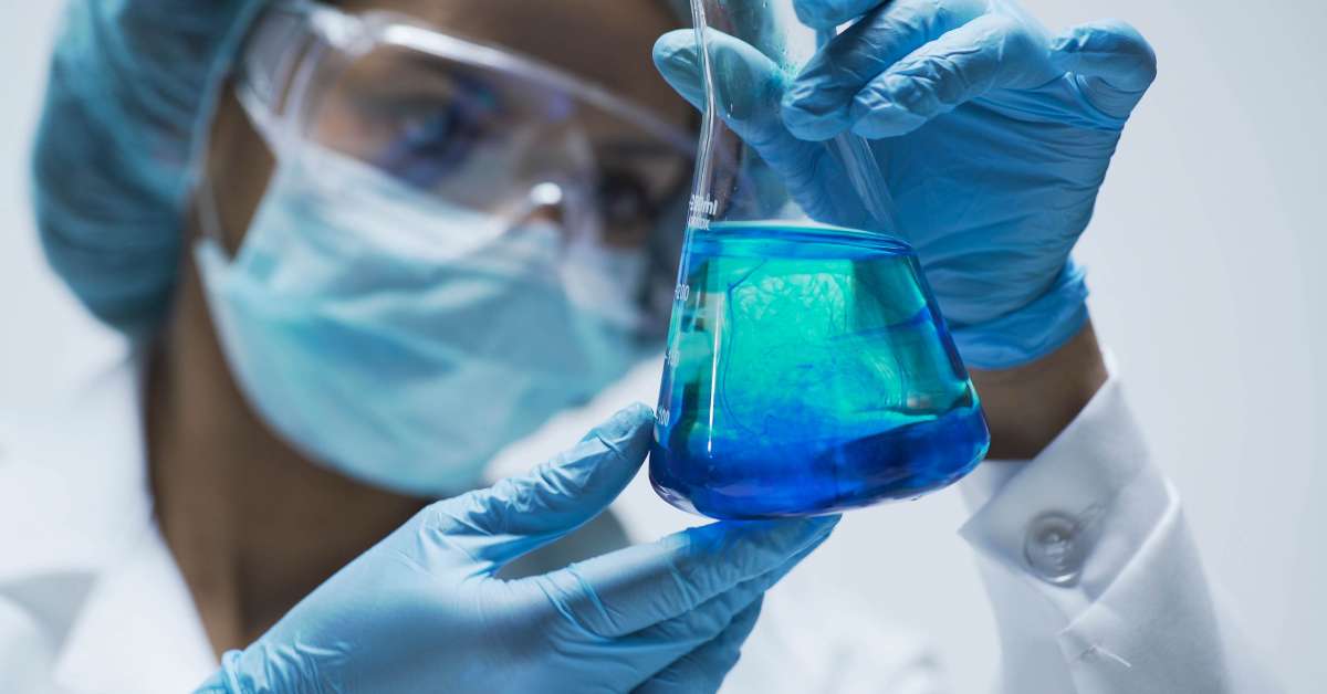 A woman holds a flask with blue liquid inside. She's wearing blue gloves, a face mask, a white lab coat, and eye glasses.