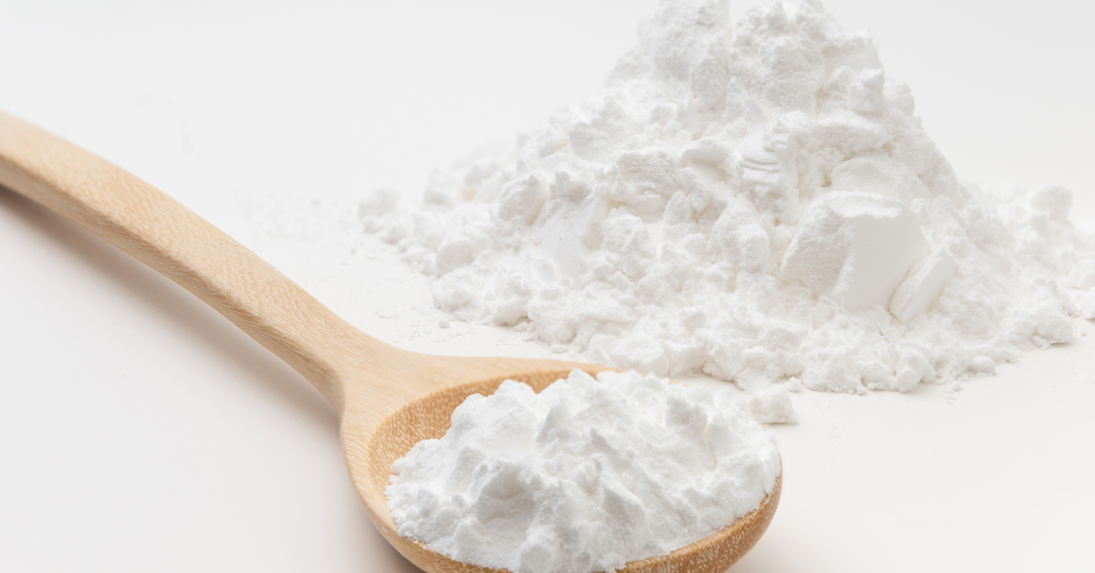 A long light brown wooden spoon filled with white powder. A pile of white powder is next to the wooden spoon.