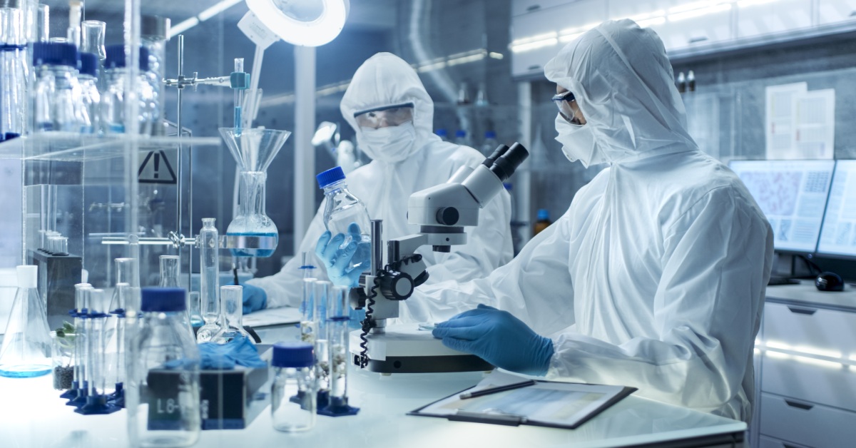 Two scientists in a laboratory wearing safety gear. They have on eye goggles, gloves, and a protective suit.