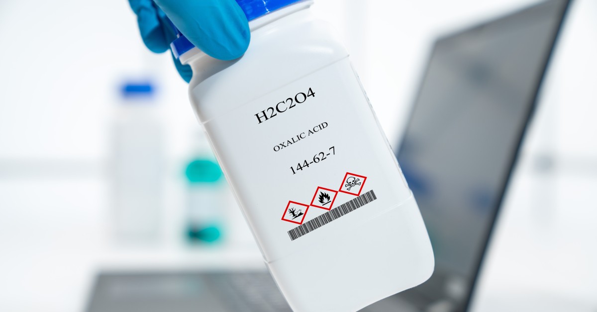 A person holds a bottle labeled "H2C2H4 OXALIC ACID 144-62-7." They are in a white laboratory near a laptop on a table.
