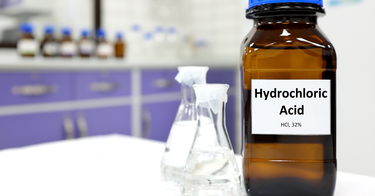 A bottle labeled hydrochloric acid that is sitting next to two smaller bottles. The bottles are inside of a laboratory.