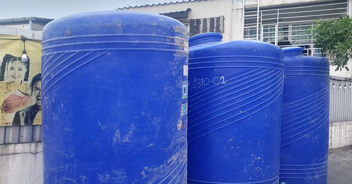 A row of blue polyethylene drums placed outside on a sidewalk. The drums are securely positioned next to a wall.