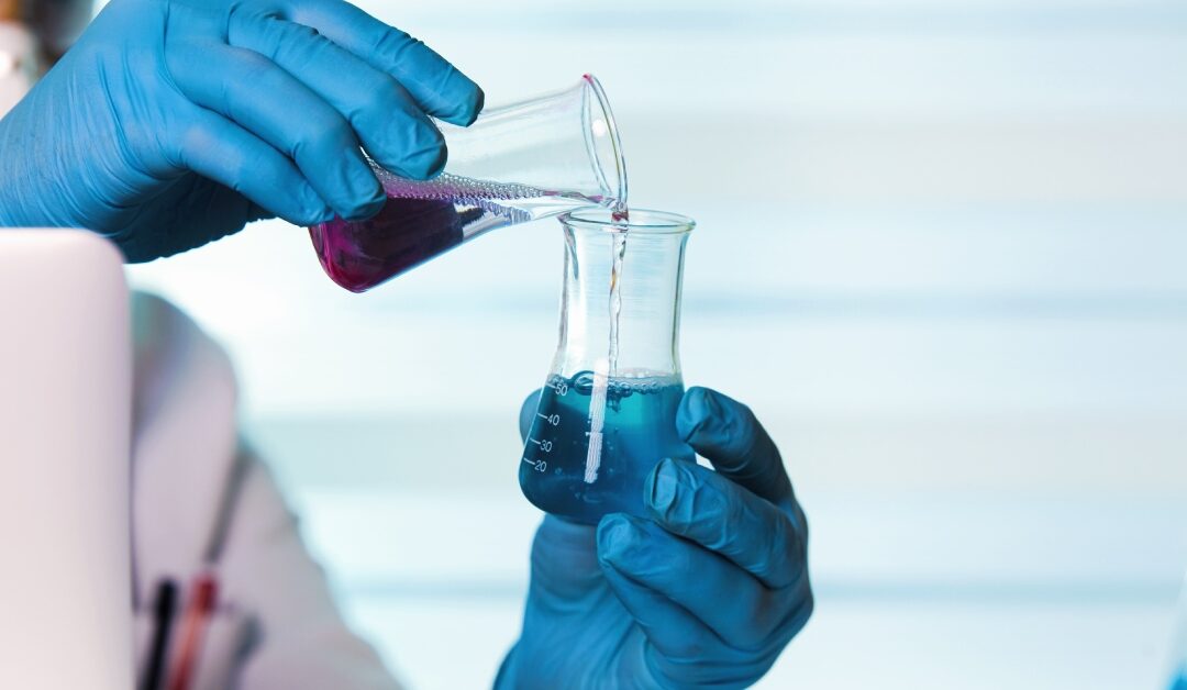 A person with gloves on pouring a glass of chemical fluid into a another glass with chemical fluid in it.