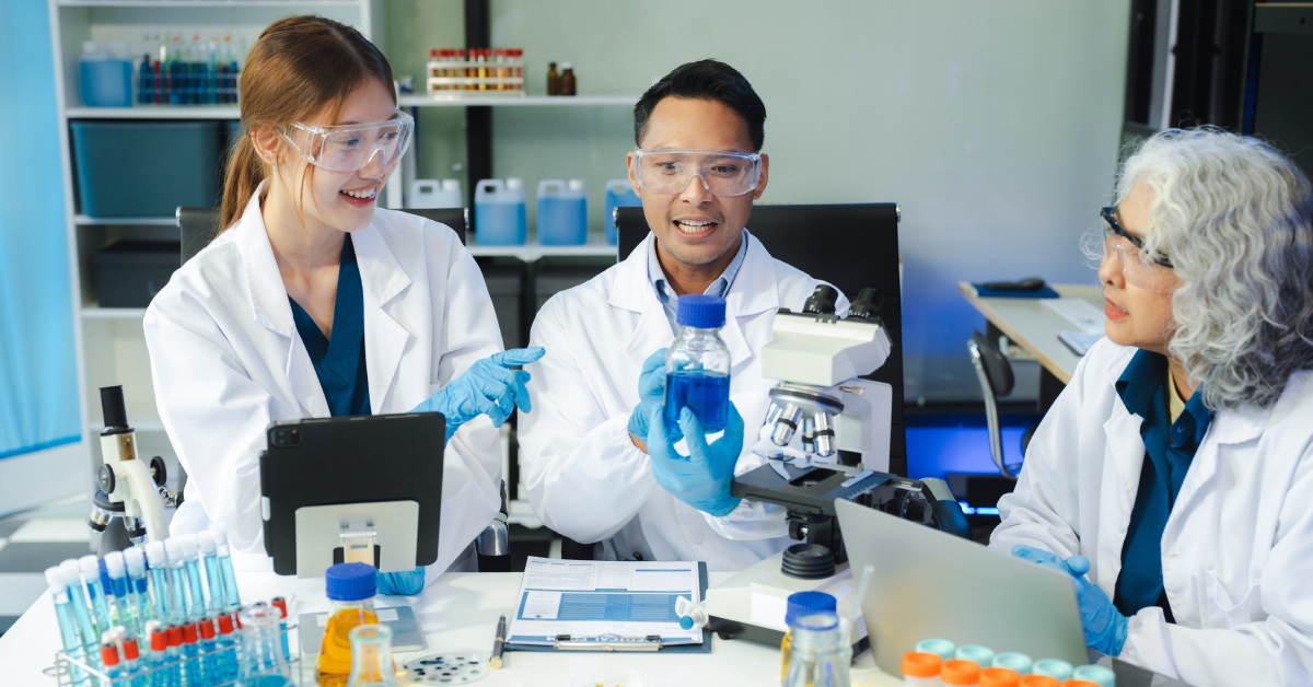 Three scientists wearing white lab coats and goggles are looking at a container with blue liquid in it.