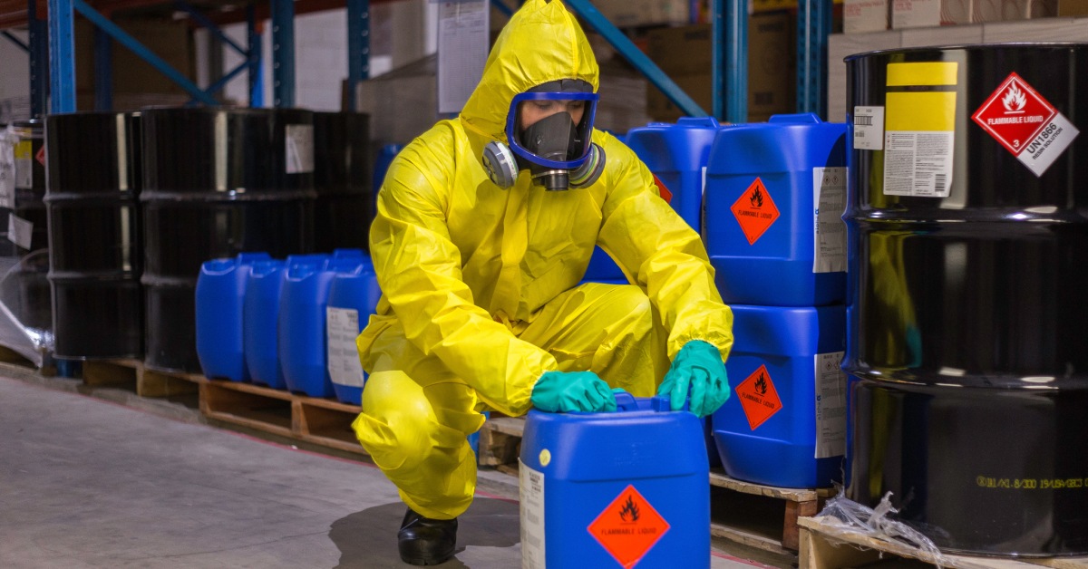 A man crouched on the floor wearing a hazmat suit. He's holding a container with the words flammable liquid on it.