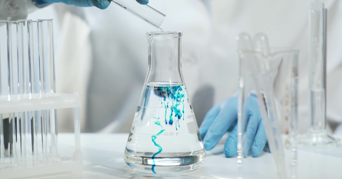 A person wearing blue gloves and a lab coat pouring liquid into a glass flask. The liquid going in the flask is turning blue.