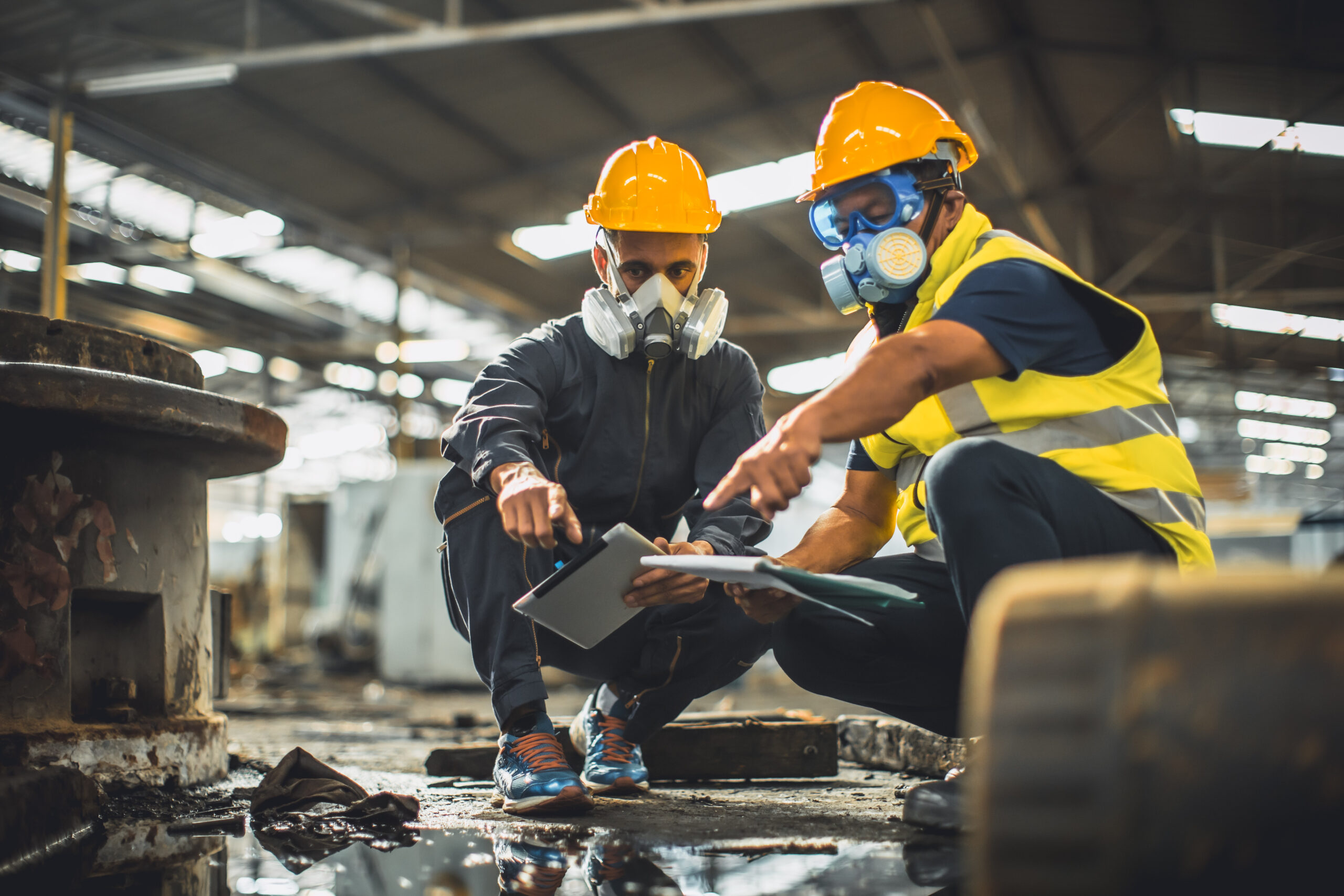 Two people crouched on the floor with helmets, goggles, and masks. They are inside of a facility.