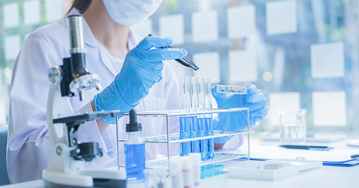 A lab worker holding a device in front of a row of test tubes. The test tubes have blue liquids in them.