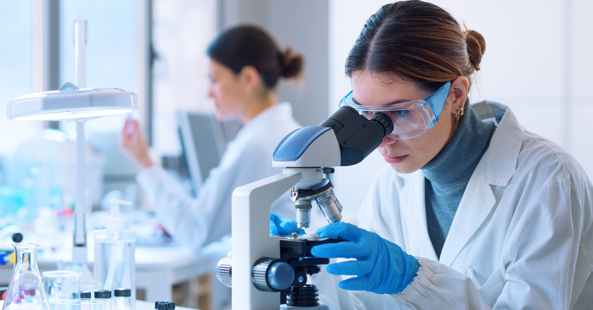 A woman looking through a microscope and wearing eyeglasses. Another woman is in the room behind her.