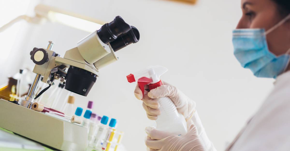 A person in a white coat spraying a white surface. They have blue gloves and a face mask on.